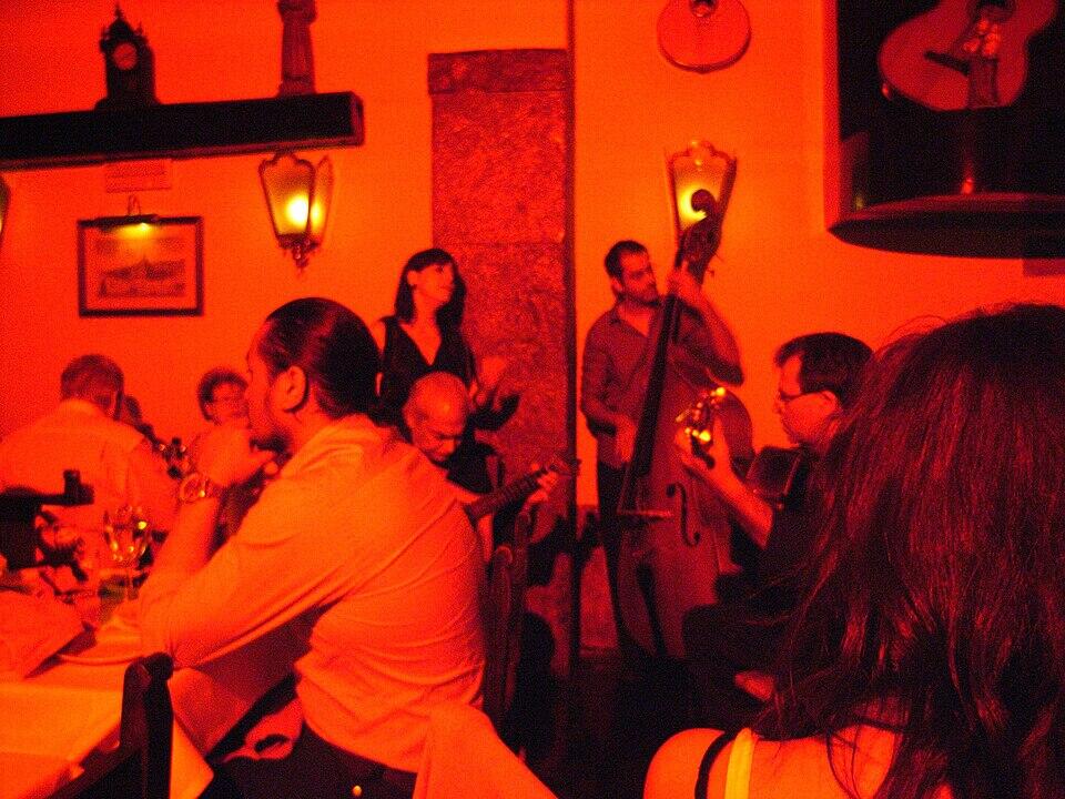 Una escena en un restaurante con poca luz en la que aparecen m&uacute;sicos tocando instrumentos, iluminados con c&aacute;lidos tonos rojos, y clientes disfrutando del ambiente.