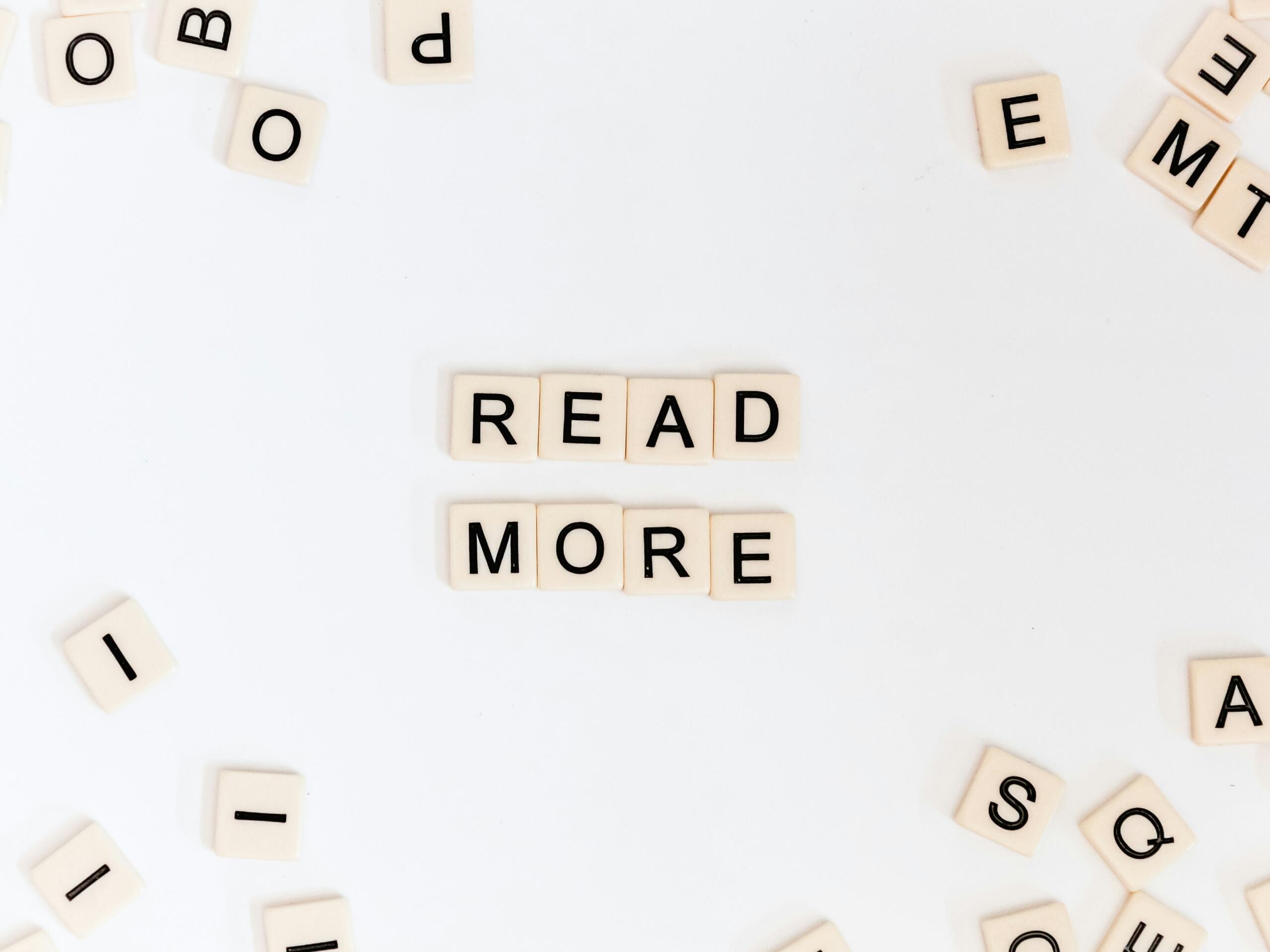 Fichas de Scrabble dispuestas para formar la frase «READ MORE» (LEER MÁS) con letras dispersas sobre una superficie blanca.